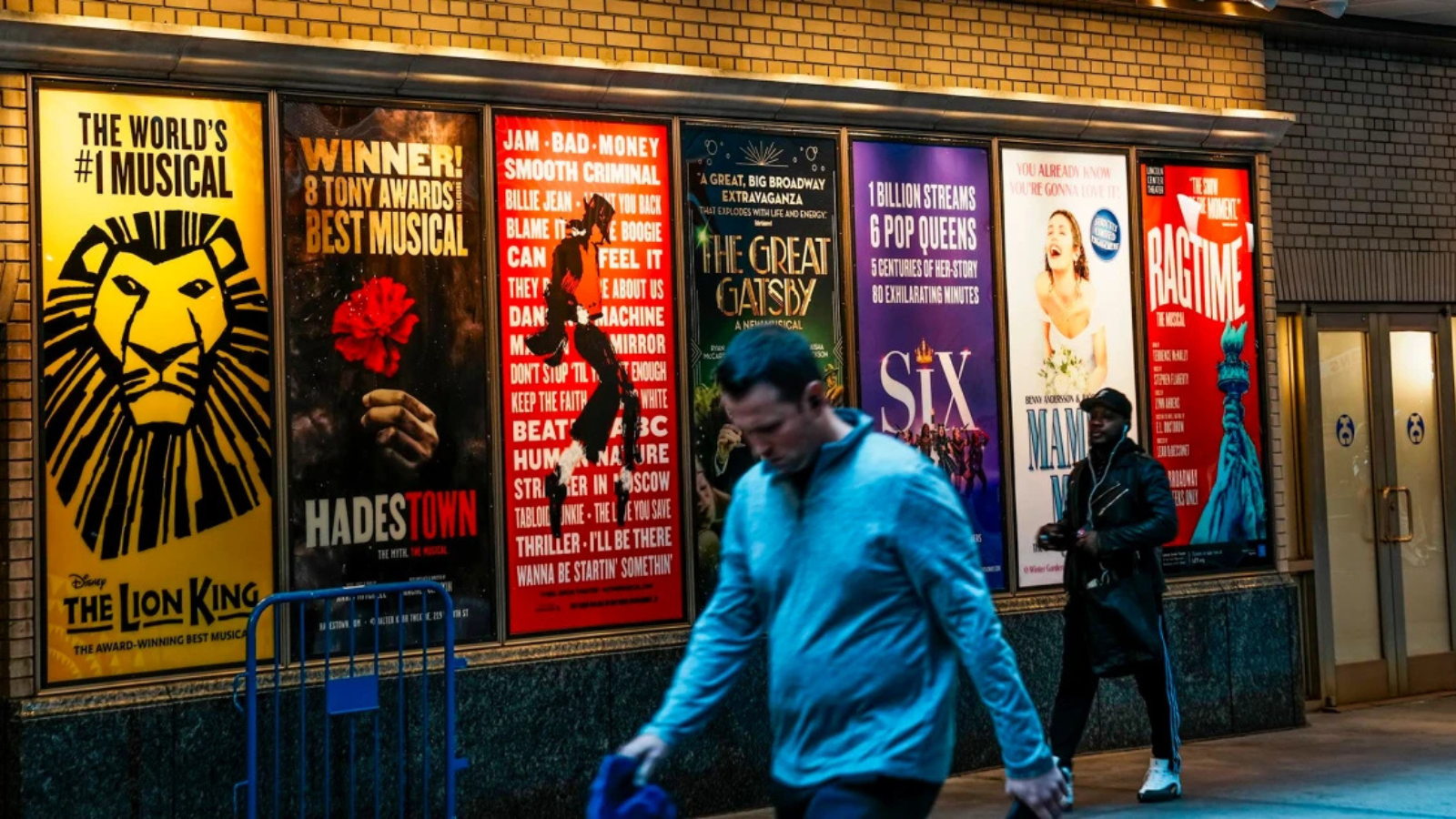 A patron walks on the streets of NYC, Broadway show posters behind them.
