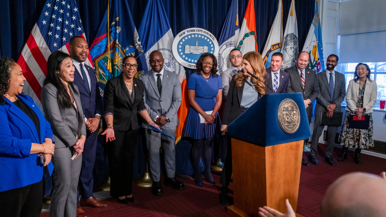 NY City Council members and Speaker Julie Menin stand together behind a podium for a press conference.