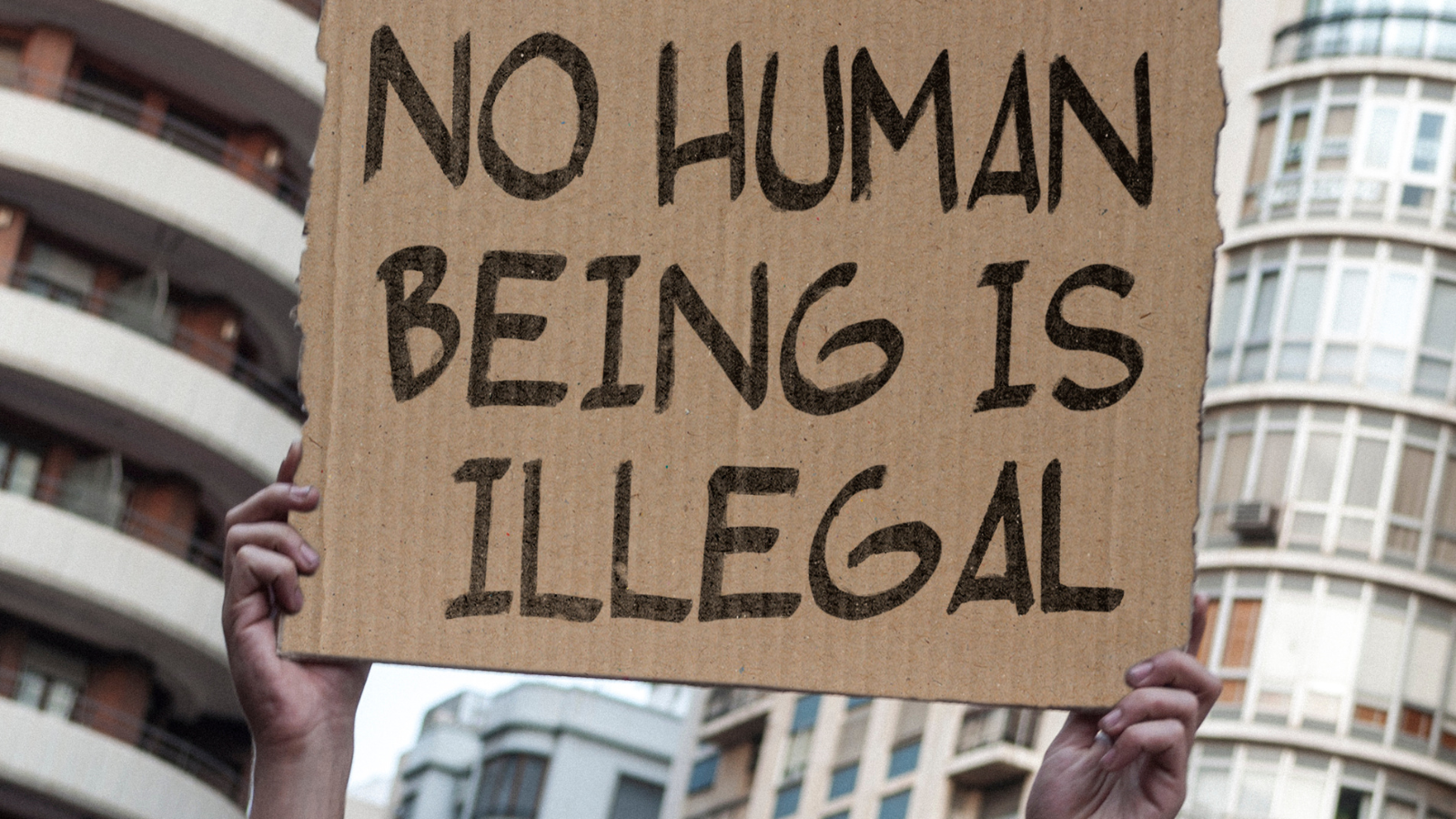 Hands holding cardboard poster that reads ‘no human being is illegal’ 