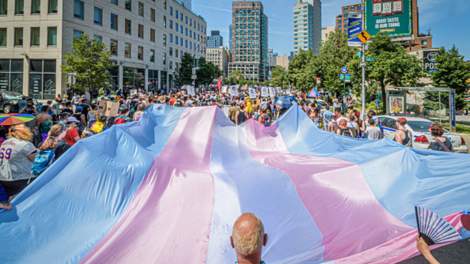 A trans flag being spread out by folks on a city street during a Pride parade