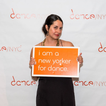 Raquel stands in front of the Dance/NYC step and repeat. She looks at the camera, wearing a black dress with a statement necklace, and her hair is pulled back. She holds an orange sign that reads, in white text, ‘i am a new yorker for dance’.